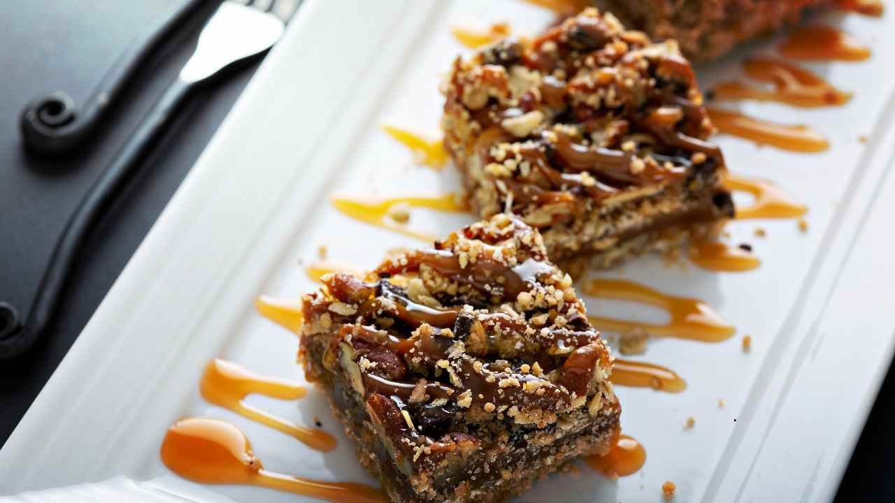 Close-up of pecan dessert bars drizzled with caramel sauce, served on a white plate with a fork in the background