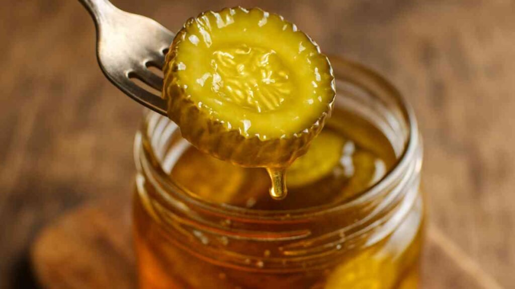Pickle slice coated in sweet syrup on a fork with jar of candied pickles in background