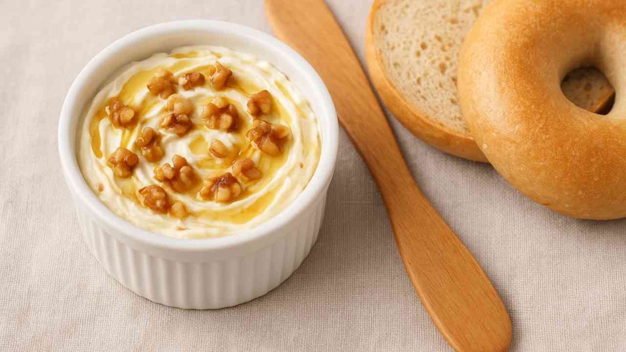 Ramekin of honey walnut cream cheese topped with chopped walnuts and honey beside a sliced bagel